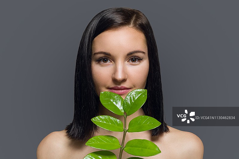 工作室美容写真：年轻深色头发女子，自然妆容，完美肌肤，灰色背景上的绿色异国情调叶子，天然化妆品、面部和身体护理产品概念图片素材
