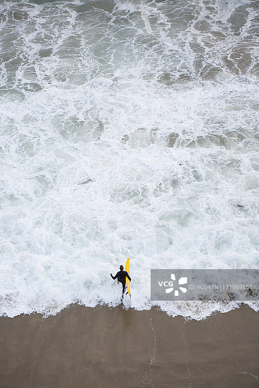 穿黑色潜水服的男子手持黄色冲浪板从上方跑入海中图片素材
