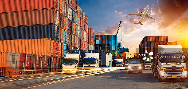 集装箱卡车在港口进行商业物流和运输，日出时船坞内有集装箱货轮和货机以及起重机桥，物流进出口和运输行业背景图片素材