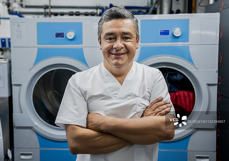 在自助洗衣店工作的英俊中年男子肖像，交叉双臂对着镜头微笑图片素材