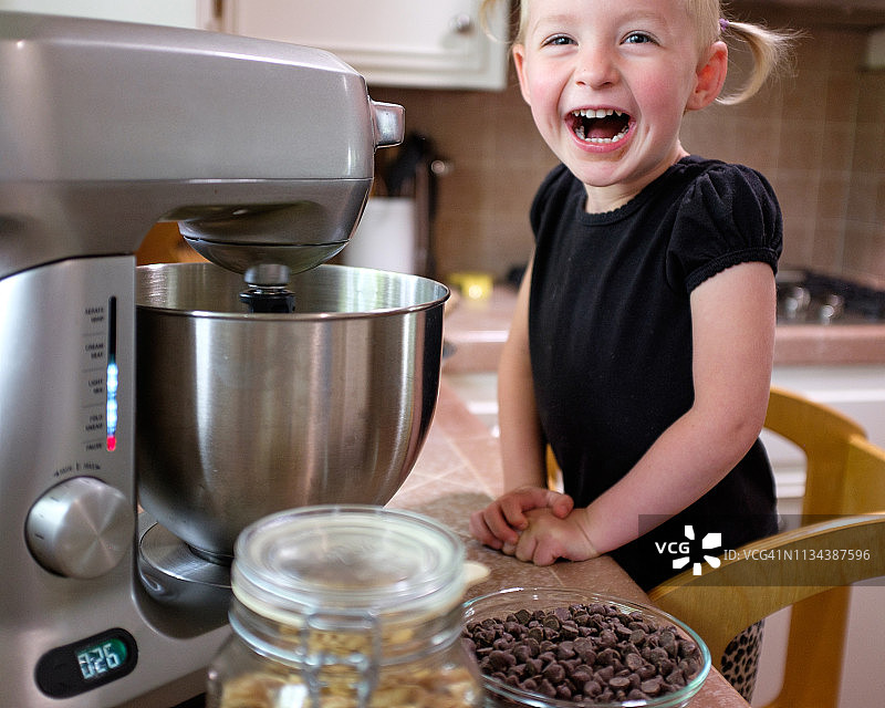 一个3岁的金发小女孩兴奋地看着，同时用电动搅拌器在家里厨房里做饼干面团图片素材