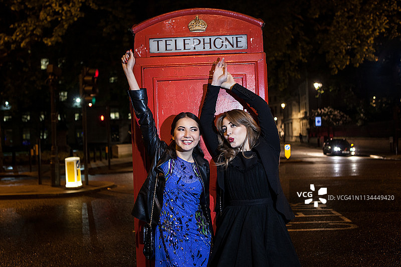 两位年轻的女性朋友在伦敦夜晚的红色电话亭前跳舞图片素材