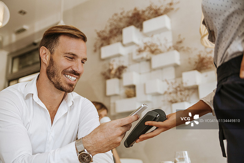 在咖啡馆用智能手机支付的微笑男人图片素材
