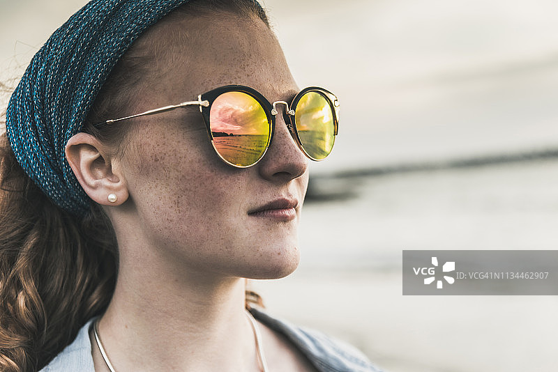 年轻女子在海滩日落时太阳镜中云朵的倒影图片素材