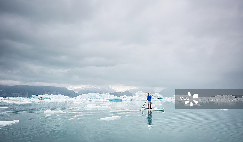 在阴沉天空下冰川泻湖上的桨板运动男子图片素材