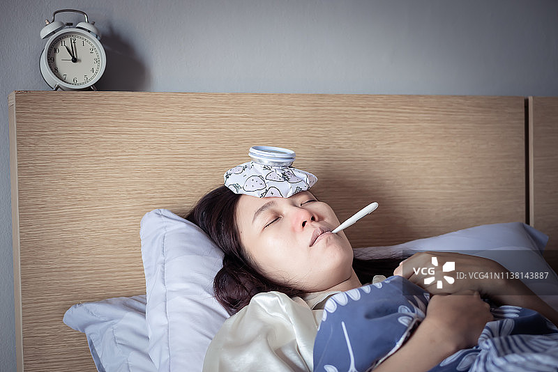 亚洲女人感冒发高烧，躺在舒适的床上用冰袋降温，并用体温计测量体温。生病在家图片素材