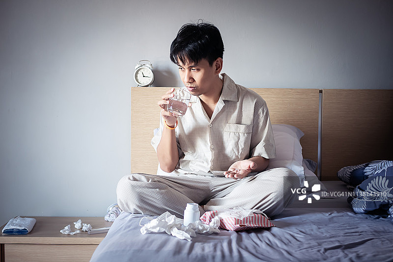 病人手拿止痛药和水杯坐在舒适的床上，旁边放着纸巾盒，在家养病图片素材
