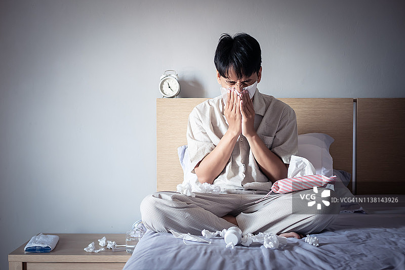 亚洲男人感冒了，坐在舒适的床上用纸巾擦鼻涕，在家生病图片素材