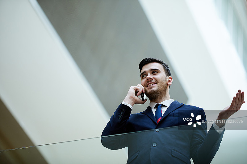 在正式套装中站在大厅里的自信的年轻男企业家在电话上讨论工作问题图片素材