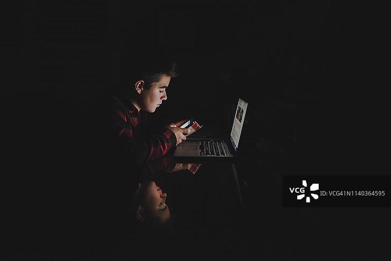 在黑暗房间里使用智能手机和笔记本电脑的青少年男孩图片素材