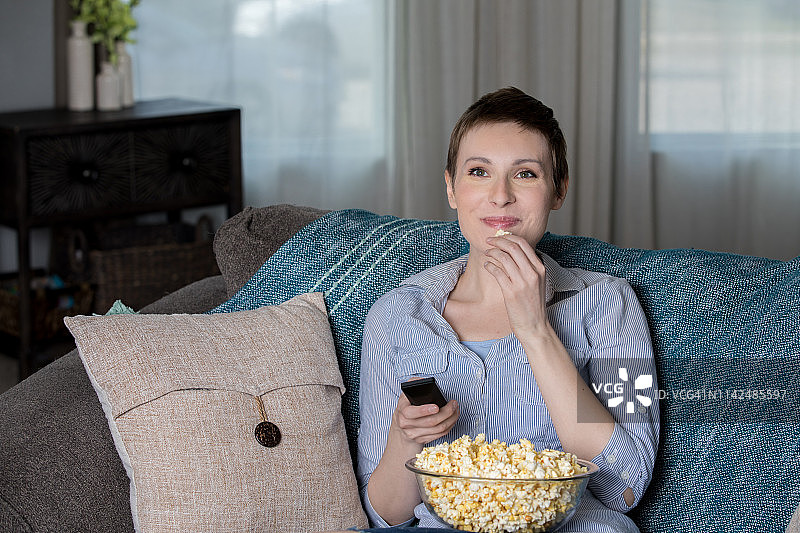 快乐的中年女人在家里的客厅里一边看电影一边吃爆米花图片素材