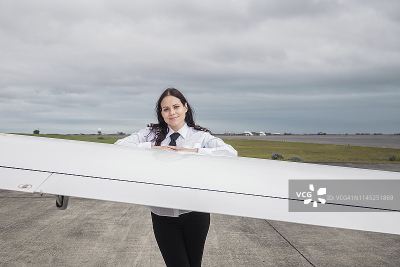 自信的女飞行员站在机场跑道上，飞机机翼旁的肖像，背景是多云的天空图片素材