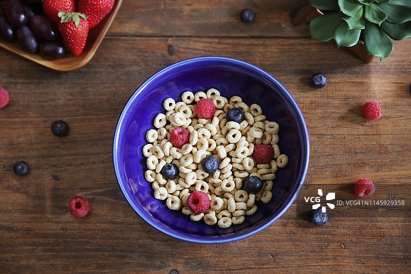 从高处拍摄的麦片碗，碗周围散落着覆盆子和蓝莓，木桌上还放着草莓和葡萄图片素材
