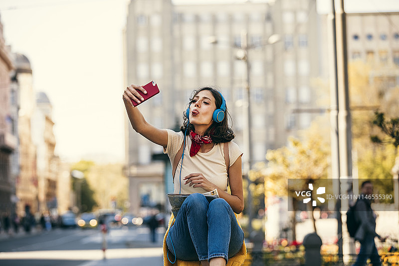有魅力的时髦女人戴着耳机，在城市里探索时听音乐和自拍图片素材