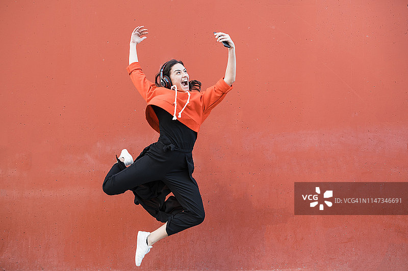 在红墙前跳跃并使用智能手机的年轻舞者图片素材
