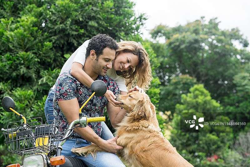 拉丁美洲男子身穿花衬衫，坐在黄色摩托车上，与金发卷发的拉丁裔女伴在摩托车上互动，并爱抚着他用后腿抱着的宠物金毛犬图片素材