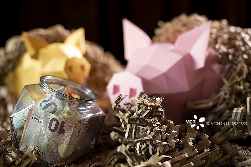 纸板“稻草”巢中的低多边形纸小猪存钱罐，带钱的低多边形玻璃罐图片素材