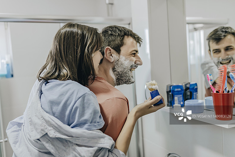 在浴室里往年轻男子脸上涂抹剃须膏的年轻女子图片素材