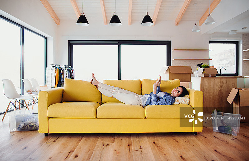 年轻女人在新家的客厅里，躺在黄色沙发上玩手机，周围都是搬家纸箱图片素材