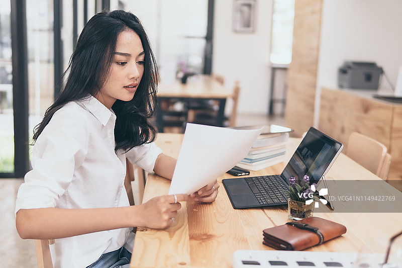 职场女性在办公桌前查看文件，使用笔记本电脑和纸质文件图片素材