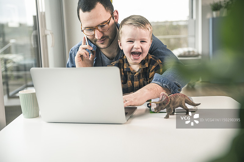父亲在家里的办公室的桌子旁工作，儿子坐在他的腿上图片素材