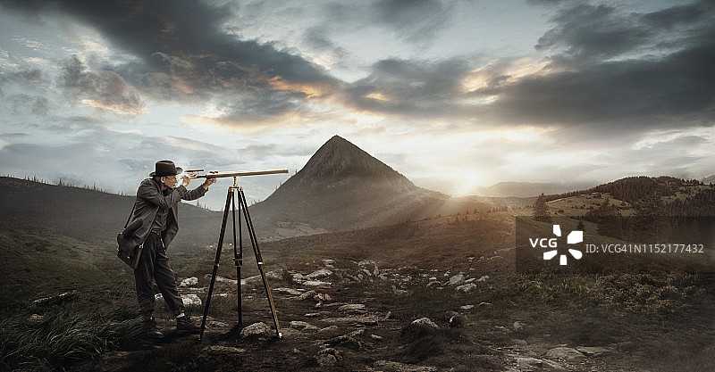 穿着经典西装戴帽子的优雅男人在山边用望远镜观看图片素材