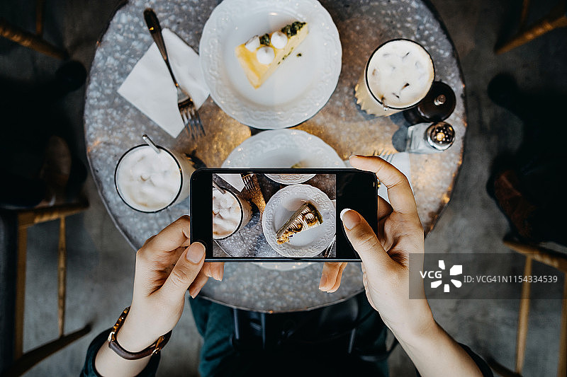 女子在咖啡馆休闲时用智能手机拍摄鲜奶油蛋糕和咖啡的照片特写图片素材