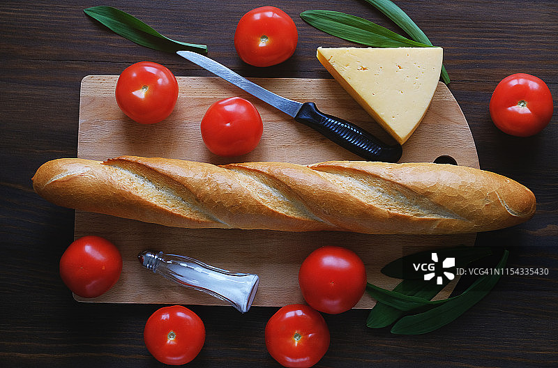 木桌上的新鲜烘焙面包、厨刀、砧板、红番茄、调味瓶、奶酪和蔬菜特写：厨房用具、烹饪和有机农场生态产品概念图片素材