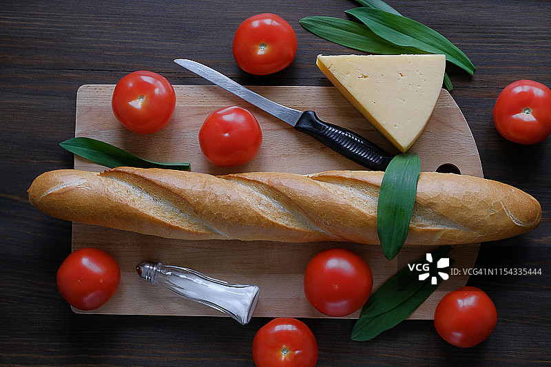 木桌上的新鲜烘焙面包、厨刀、砧板、红番茄、调味瓶、奶酪和蔬菜特写：厨房用具、烹饪和有机农场生态产品概念图片素材