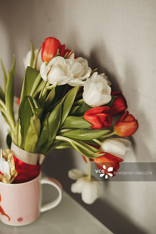 白色桌子上，浅色墙壁背景的马克杯中装着新鲜的春季白色和红色郁金香。母亲节、情人节、3月8日、生日、婚礼花束。选择性聚焦。图片素材