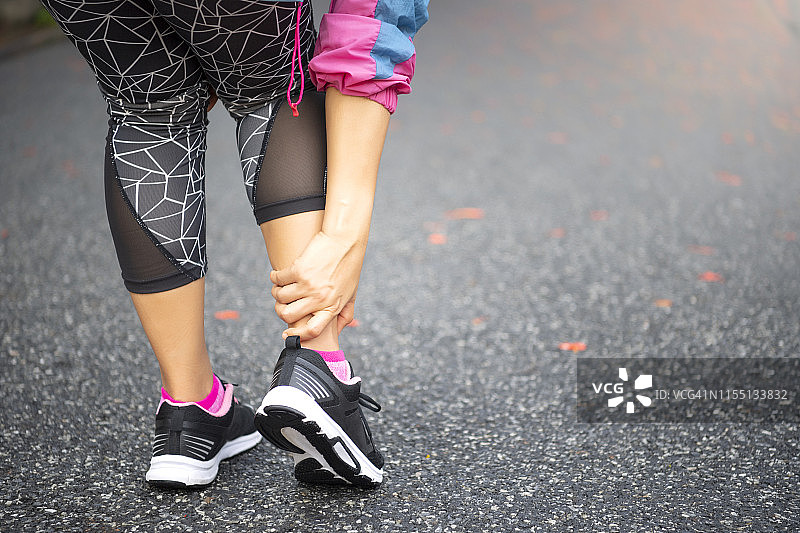 亚洲女性跑步后站在路上，手扶受伤的脚踝图片素材
