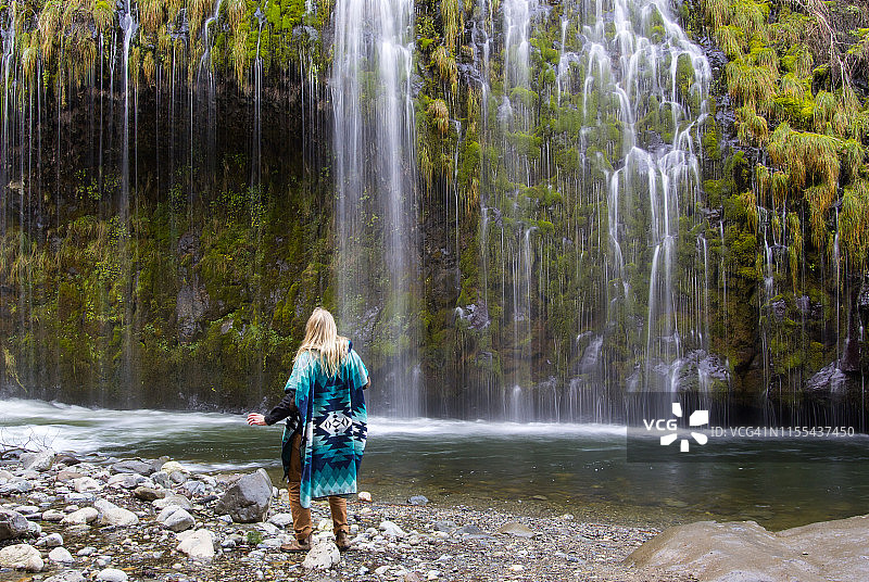 男人穿着蓝绿色披风站在瀑布和宁静的森林前图片素材