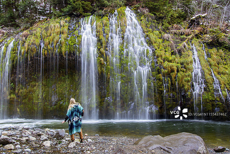 男人穿着蓝绿色披风站在瀑布和宁静的森林前图片素材