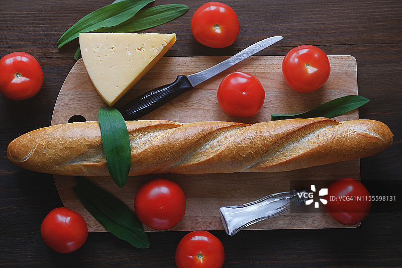 木桌上的新鲜烘焙面包、厨刀、砧板、红番茄、调味瓶、奶酪和蔬菜特写：厨房用具、烹饪和有机农场生态产品概念图片素材