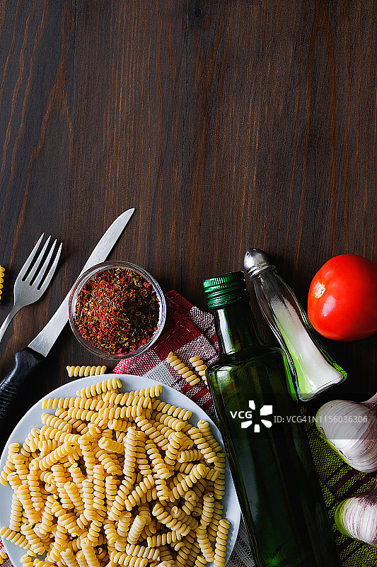意大利面、橄榄油、香料、西红柿、盐、大蒜、刀叉摆放在深色木桌上。顶部视角，留有文案空间。意大利美食概念。烹饪素食或纯素食品。图片素材