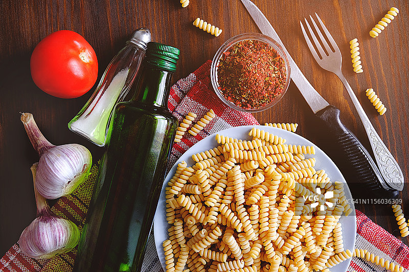 意大利面、橄榄油、香料、西红柿、盐、大蒜、刀叉摆放在深色木桌上。顶部视角，留有文案空间。意大利美食概念。烹饪素食或纯素食品。图片素材