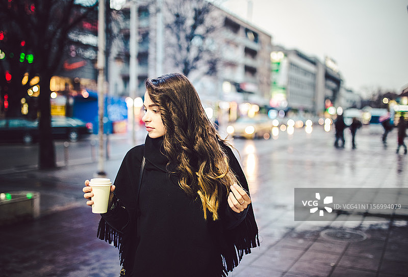 一位迷人的年轻女子在城市里散步，手里拿着一杯热咖啡图片素材