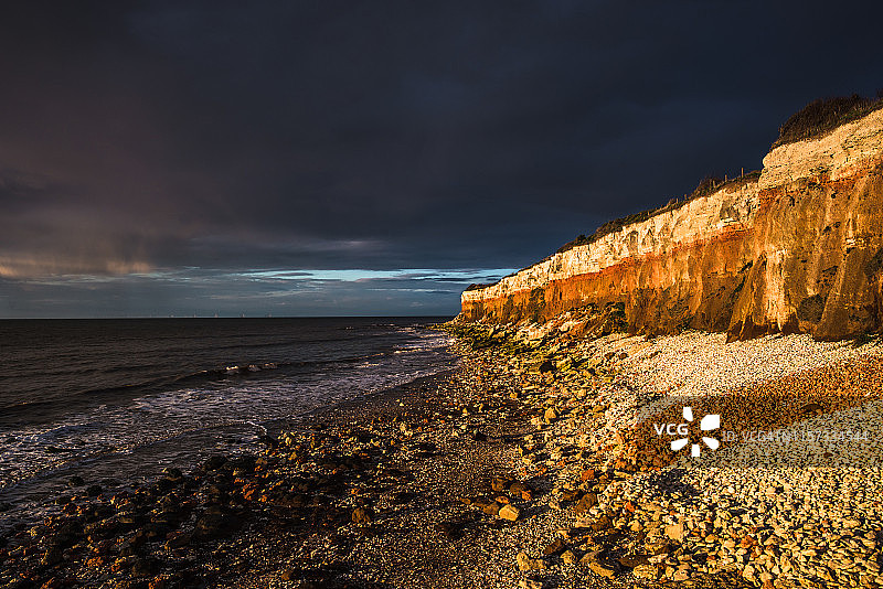 亨斯坦顿悬崖在日落时分，天空阴暗多云，位于诺福克海岸，白色白垩覆盖着红色石灰石，形成色彩鲜艳的地貌。英格兰，英国。图片素材