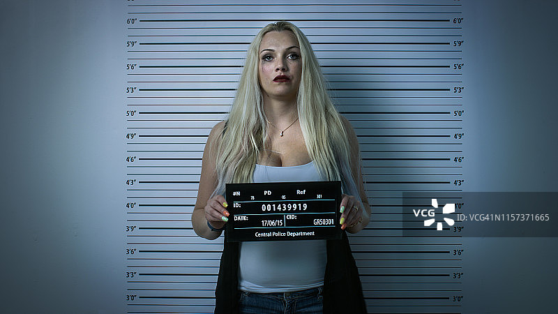 在警察局，被捕女子拍摄正面档案照，衣着暴露，浓妆艳抹，头发凌乱，手持标语牌，背景是身高表，小插图滤镜图片素材