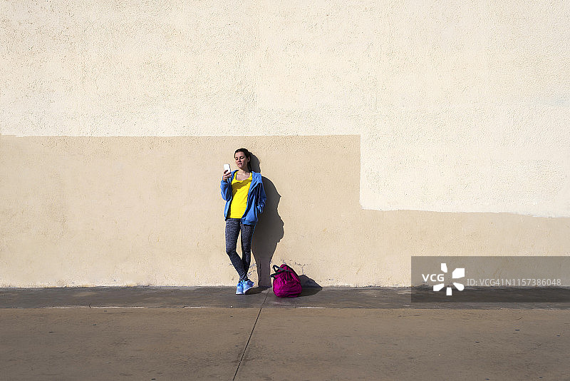 身穿运动服，靠在墙上使用手机的年轻健康女性图片素材