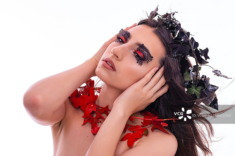 有魅力的年轻女模特肖像。女孩在白色背景的工作室里，头上戴着装饰品摆姿势。眼睛上化着奇特的妆容，像黑色和红色的叶子一样的装饰。图片素材