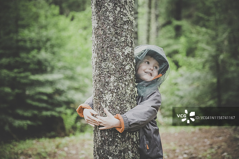 拥抱树木戴着蚊帐帽的小红发男孩图片素材