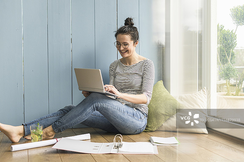 笑容满面的女人坐在家里的窗边，使用笔记本电脑和文件夹工作图片素材