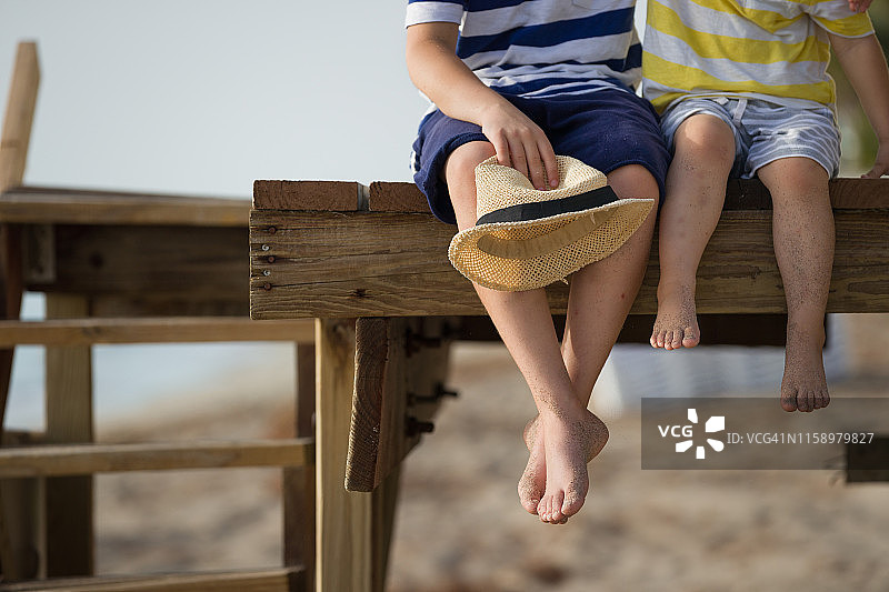 孩子们坐在木制码头上享受海滩夏日。男孩赤脚。海边度假。户外。兄弟姐妹。海边的兄弟。图片素材
