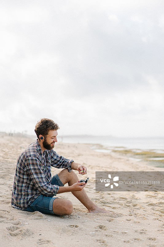 男人坐在海滩上，戴着无线耳机听音乐图片素材