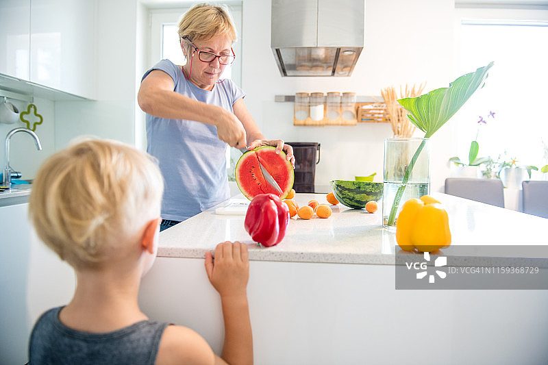 新厨房里的年长妇女正在为一个金发小男孩切西瓜图片素材