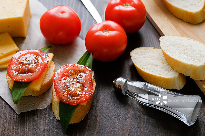 木桌上的面包片、三明治、刀、砧板、红番茄、盐瓶、奶酪和蔬菜特写。烹饪早餐或零食，有机农场产品，居家生活图片素材