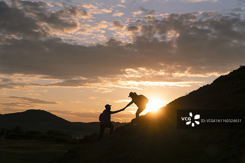 日落时分，在多云背景下，朋友互相帮助登上山顶的剪影图片素材