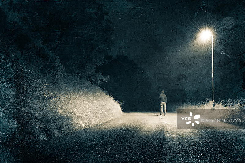 一个孤独的蒙面人站在乡村公路的中央，在阴森恐怖的夜晚的路灯下图片素材