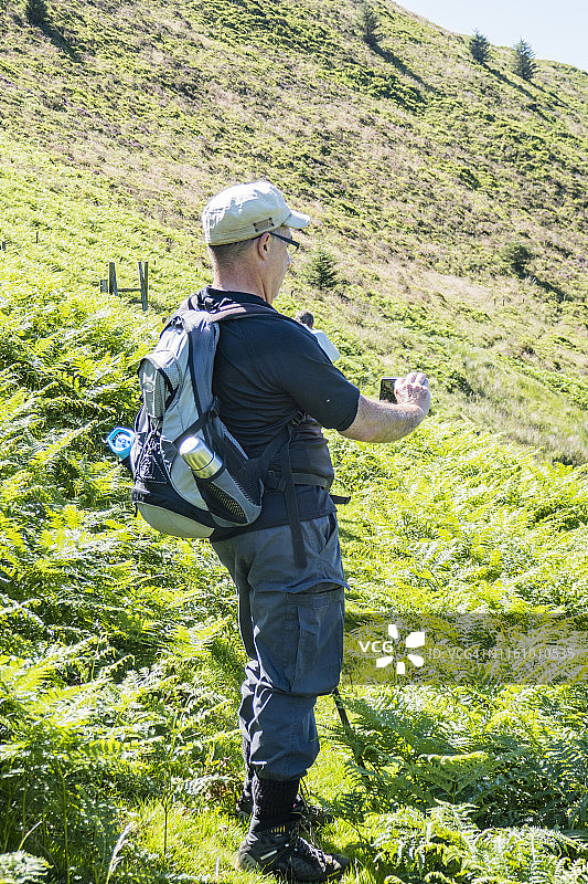 年长的白人男子在英国威尔士迪纳斯·马德威和布尔赫·林·巴赫之间的寒武纪小路上用手机拍照（7月）图片素材